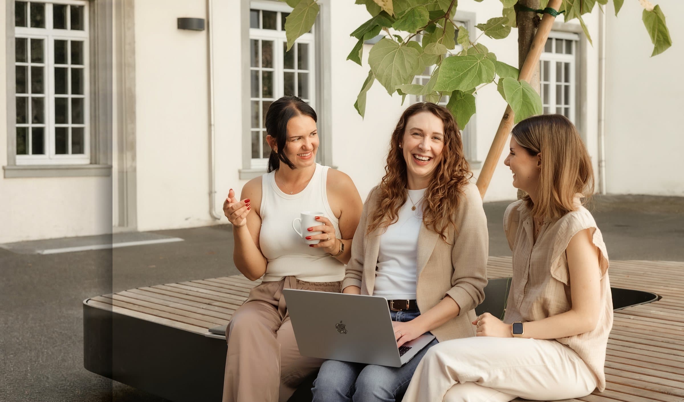 Drei Kolleginnen lachen mit Kaffee und Laptop in der Pause im sonnigen Innenhof